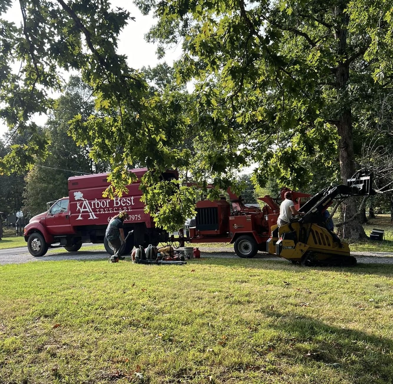 Tree Service Two workers stand near a red Arbor Best tree care truck and a yellow skid steer under a large tree, with tools and equipment spread on the grass—a sunny day showcasing professional arborist services. Middle Tennesseer